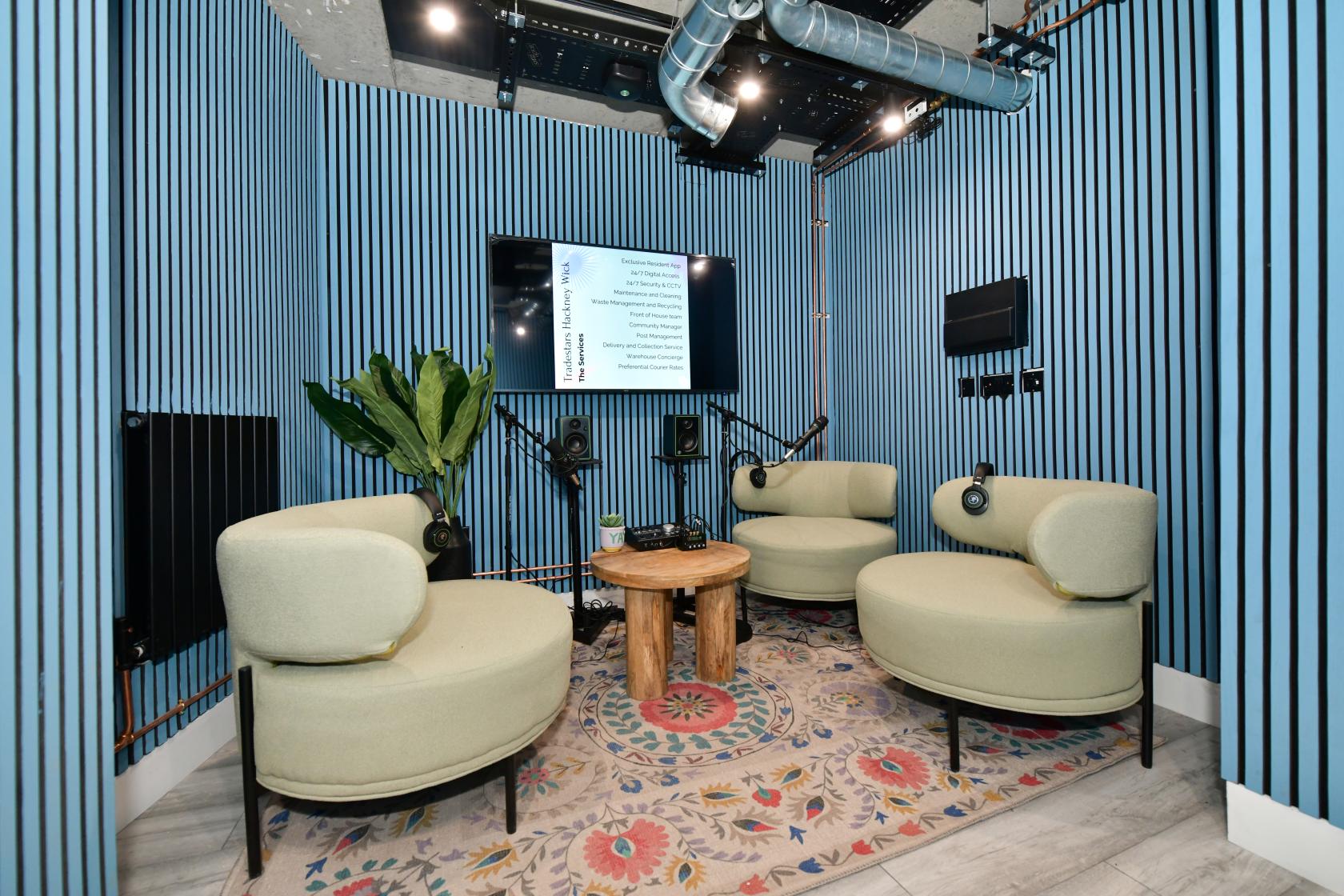 A podcast studio with curved armchairs, microphones, a round wooden table, wall-mounted screens, and a patterned rug at Tradestars Hackney Wick - Bookable Spaces.