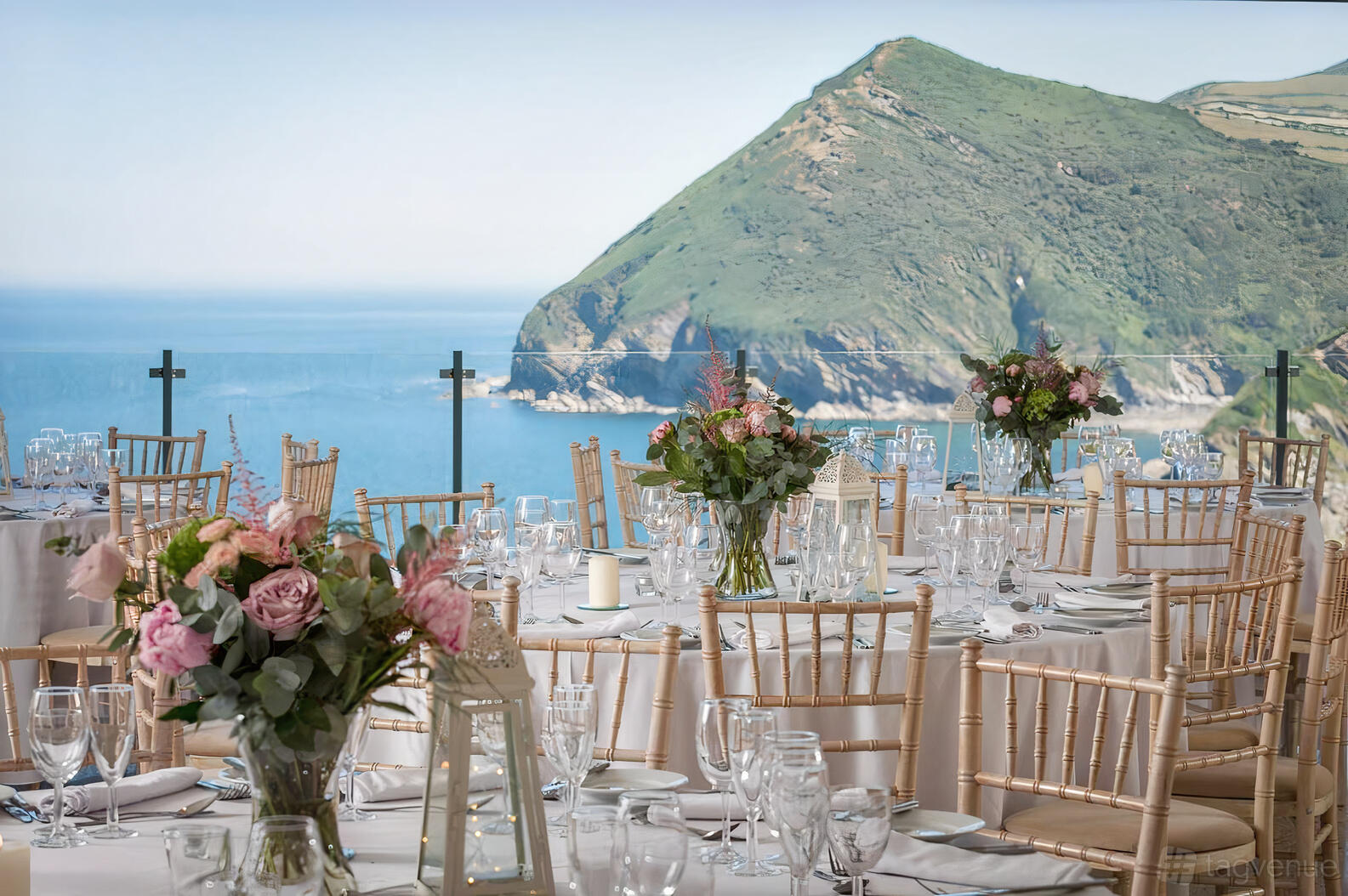 A function room with round tables set with glassware, floral centerpieces, and coastal views at The Venue At Sandy Cove.