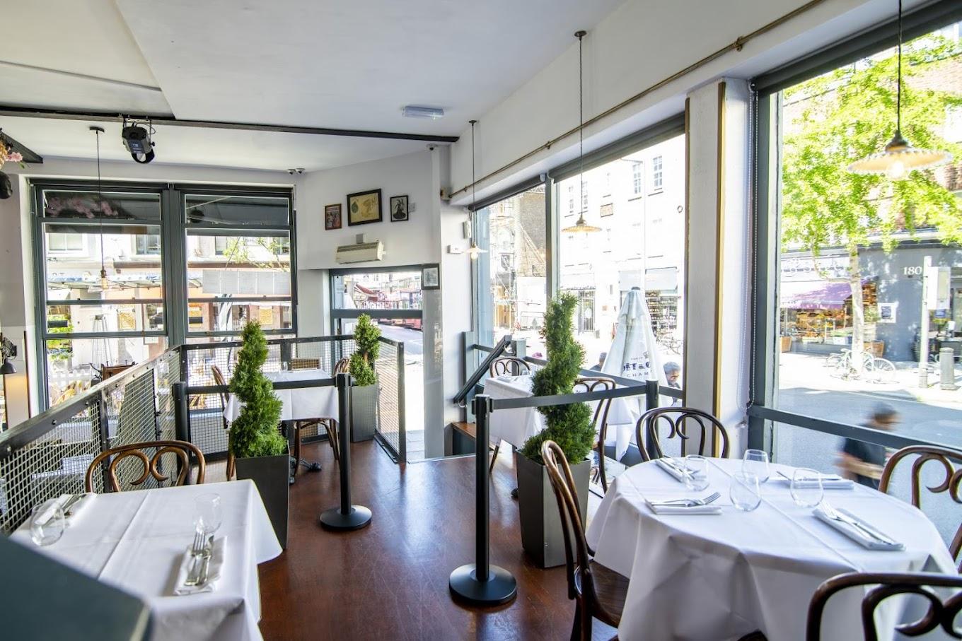 An event space in a restaurant with large windows, round tables with white linens, and street views at The Goat Chelsea.