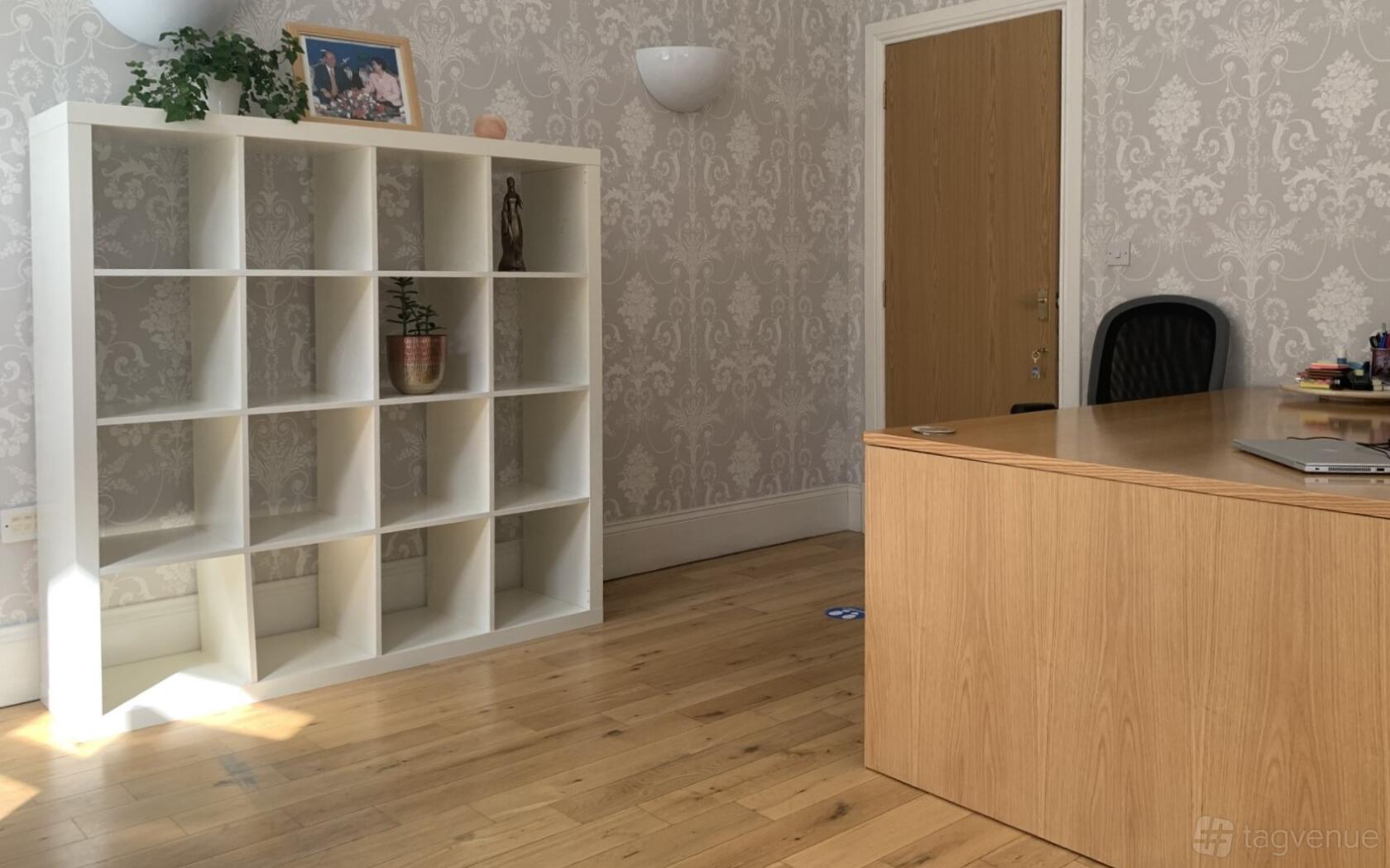 An office space with a wooden desk, open white shelving, potted plants, and patterned wallpaper at Peace Embassy FFWPU Scotland.