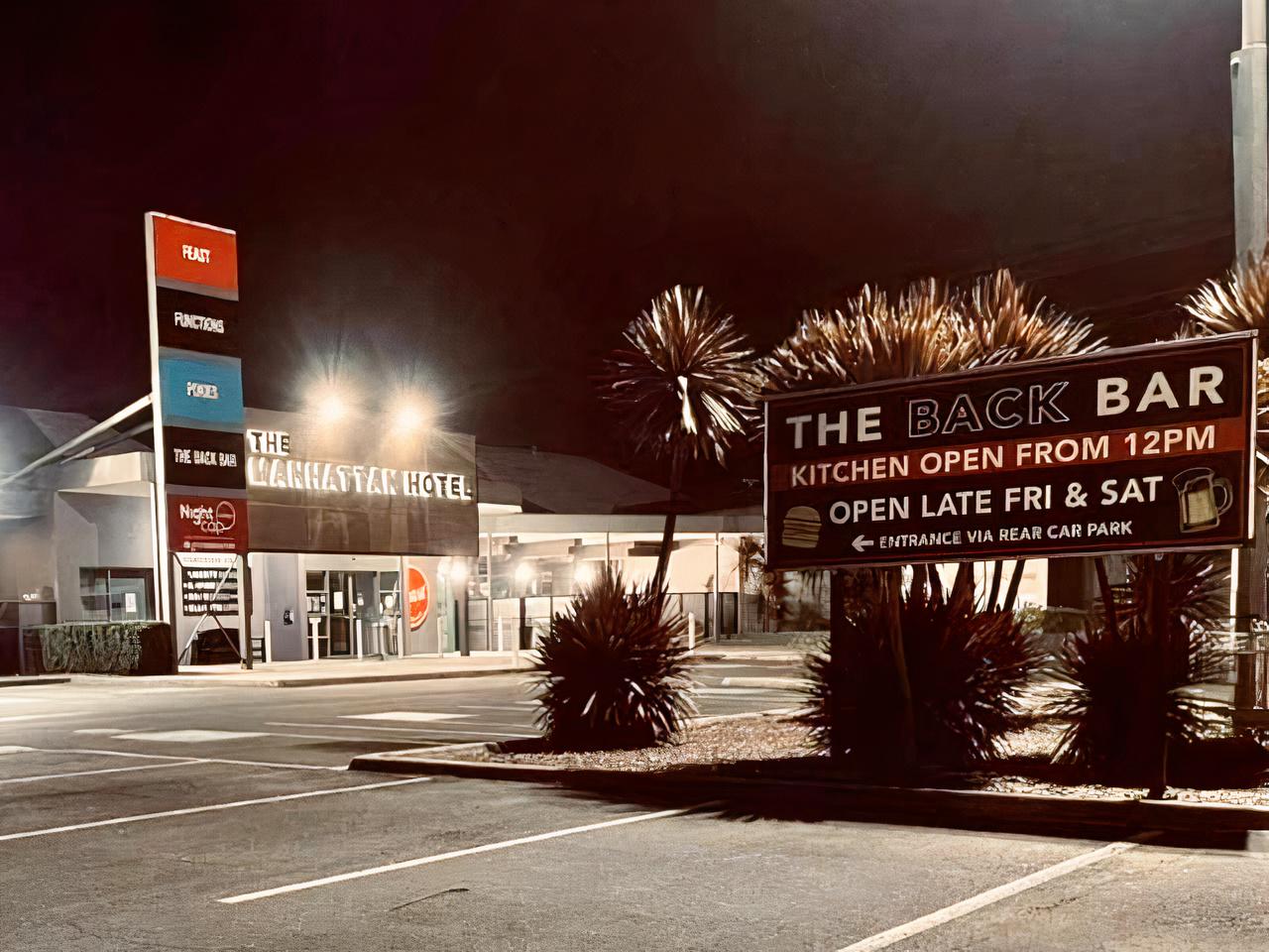A pub exterior with illuminated signage, palm trees, and a parking lot at Manhattan Hotel.