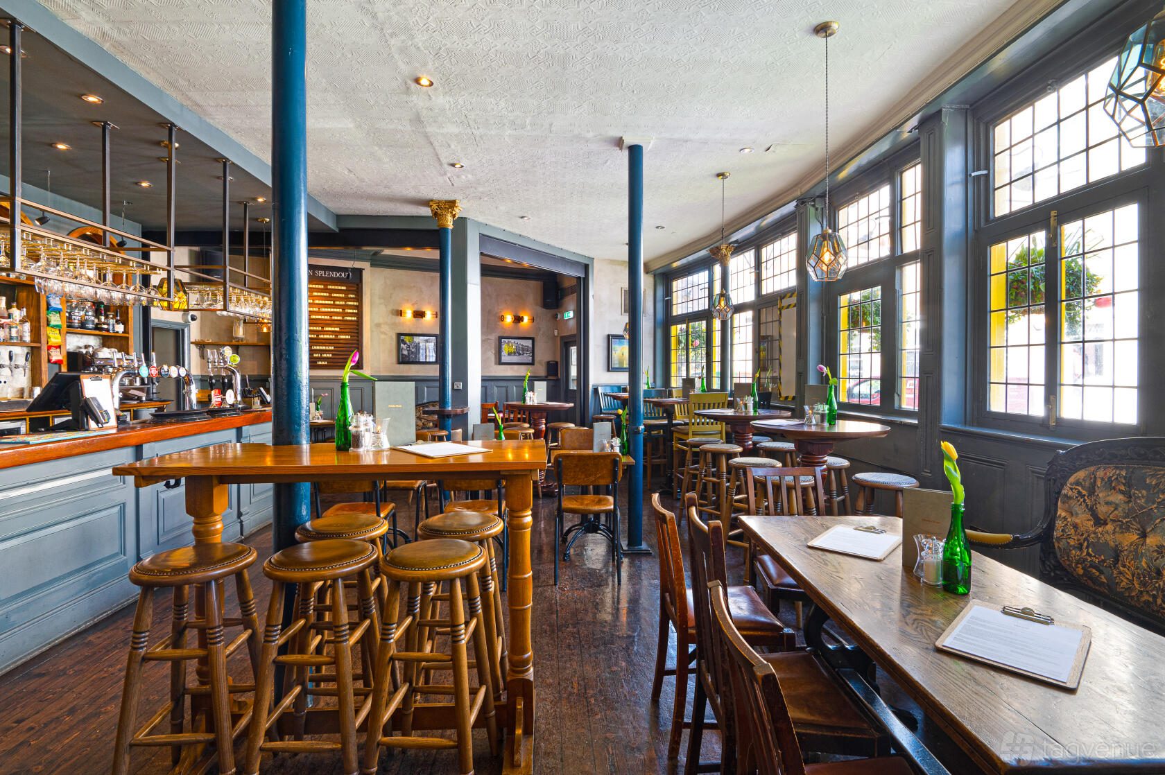 A pub with wooden tables and stools, large leaded glass windows, and a long bar at The Sun In Splendour.