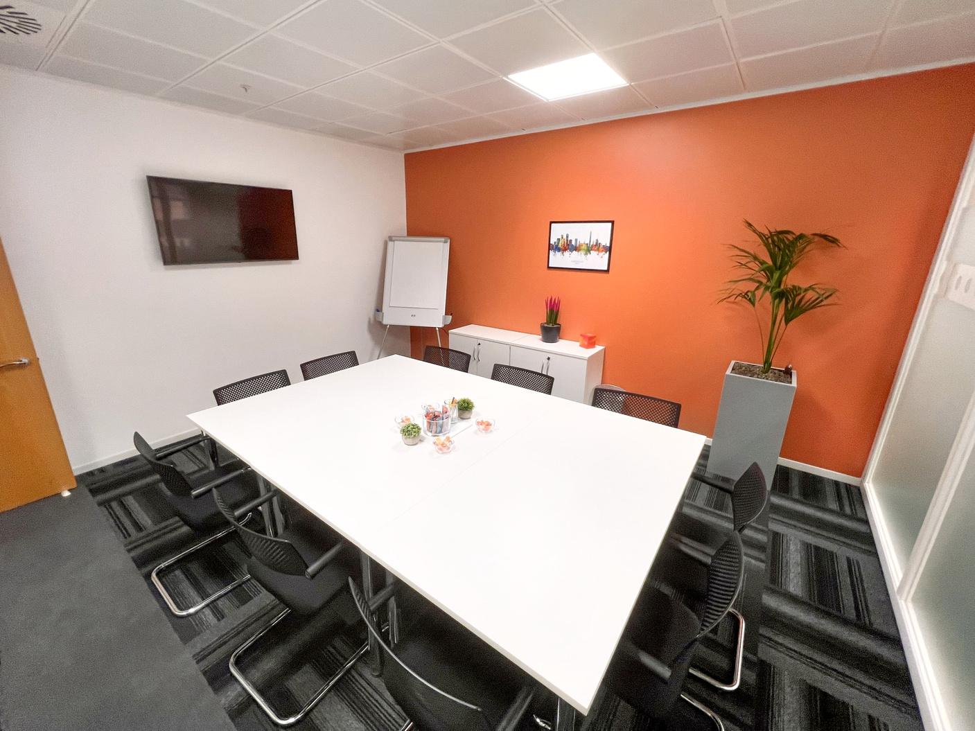 A meeting room with a white table, black chairs, orange accent wall, and wall-mounted TV at Eleven Brindleyplace.
