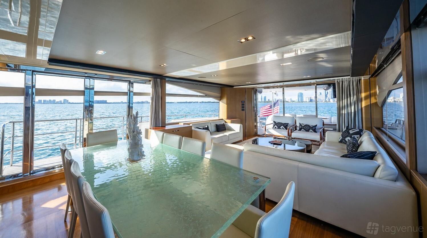 A boat lounge with glass dining table, white leather sofas, and large windows overlooking the water at Luxe Charters.