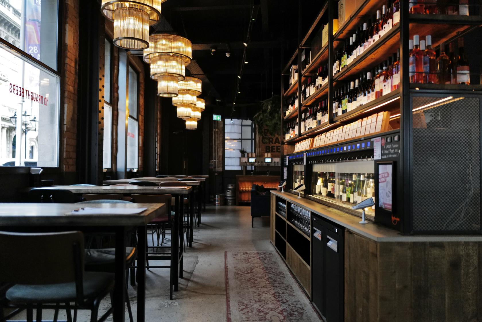 An event space in a bar with high tables, hanging chandeliers, exposed brick walls, and a self-serve wine station at Vagabond Birmingham.
