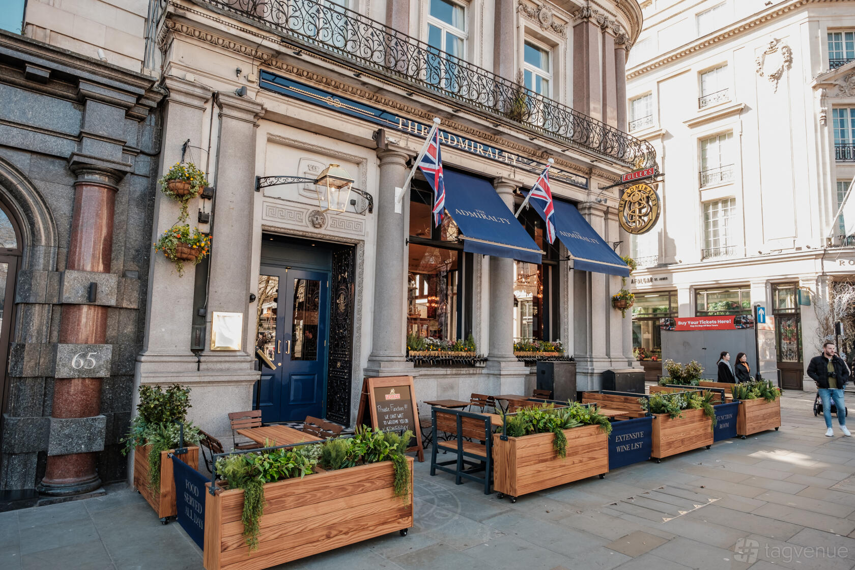A pub with outdoor wooden seating, blue awnings, and hanging flower baskets at The Admiralty.