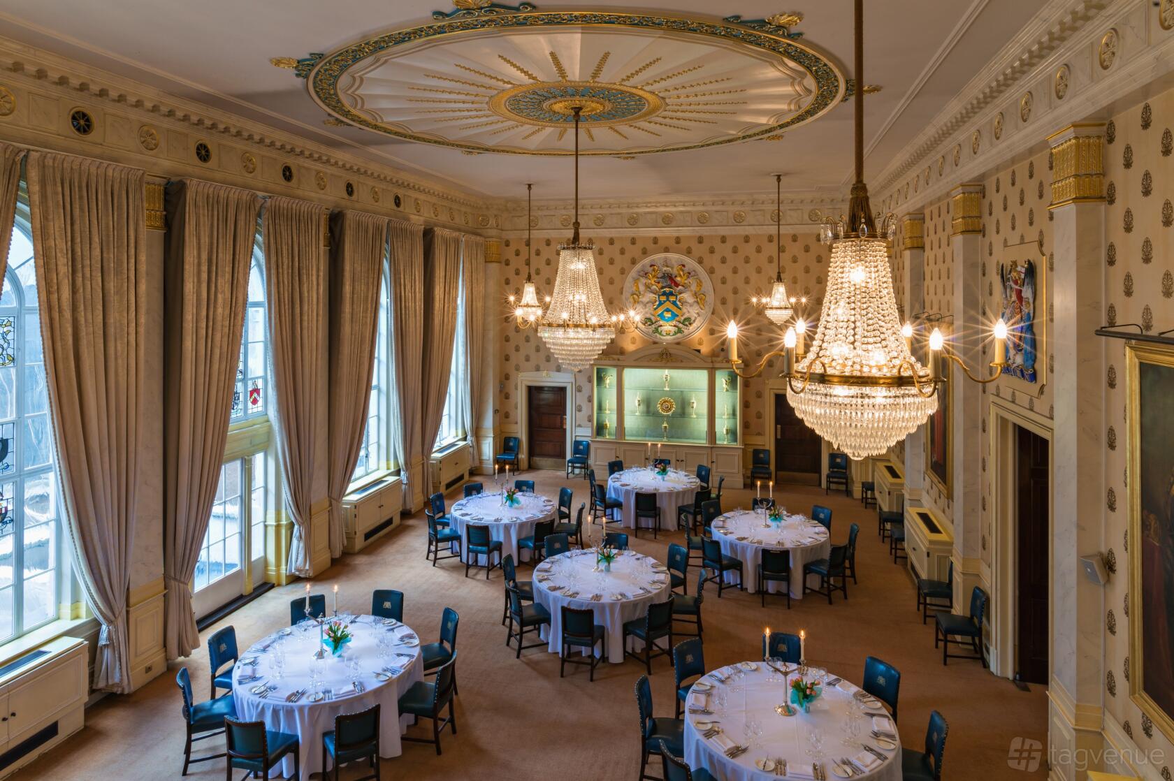 A grand hall with high ceilings, large arched windows, chandeliers, and round tables set for dining at Saddlers’ Hall.