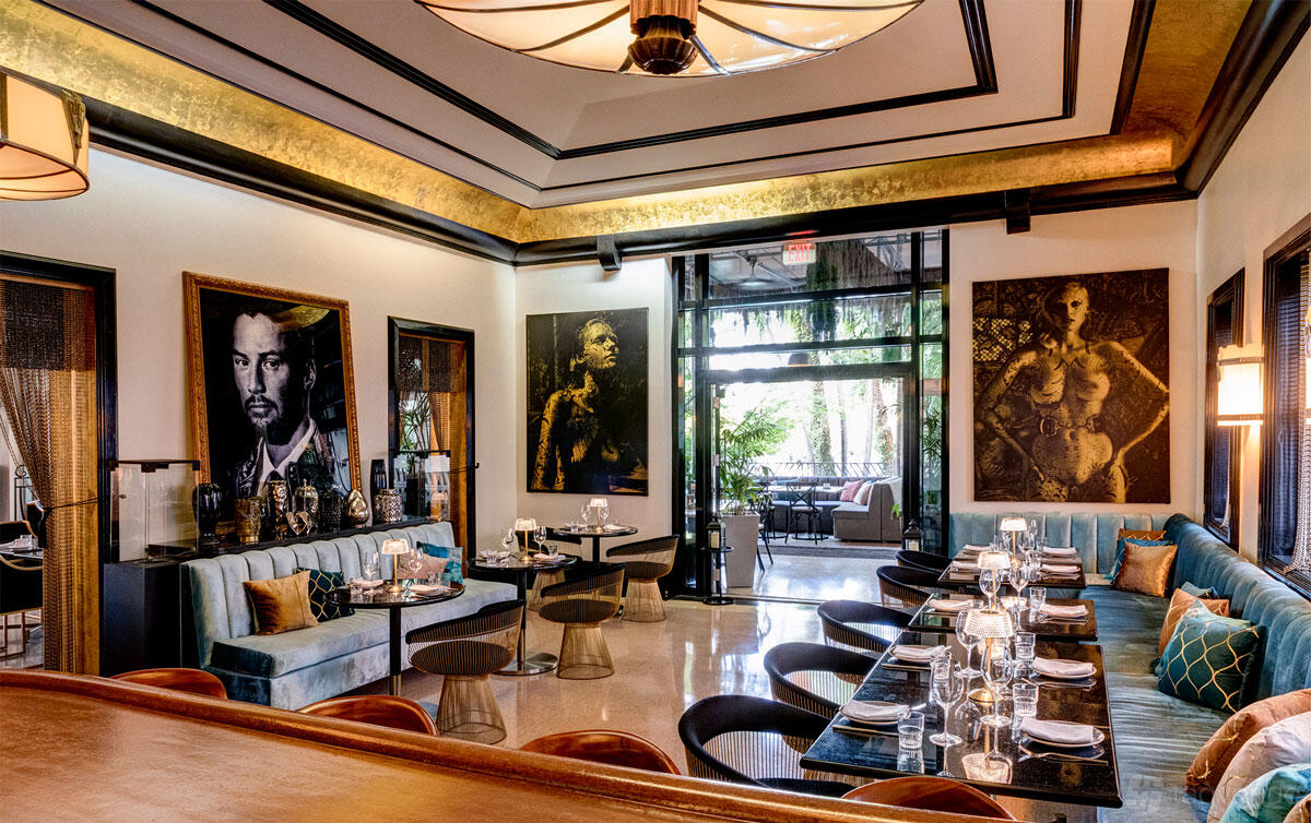A hotel bar with velvet seating, round tables, large portrait art, and glass doors opening to greenery at National Hotel Miami Beach.