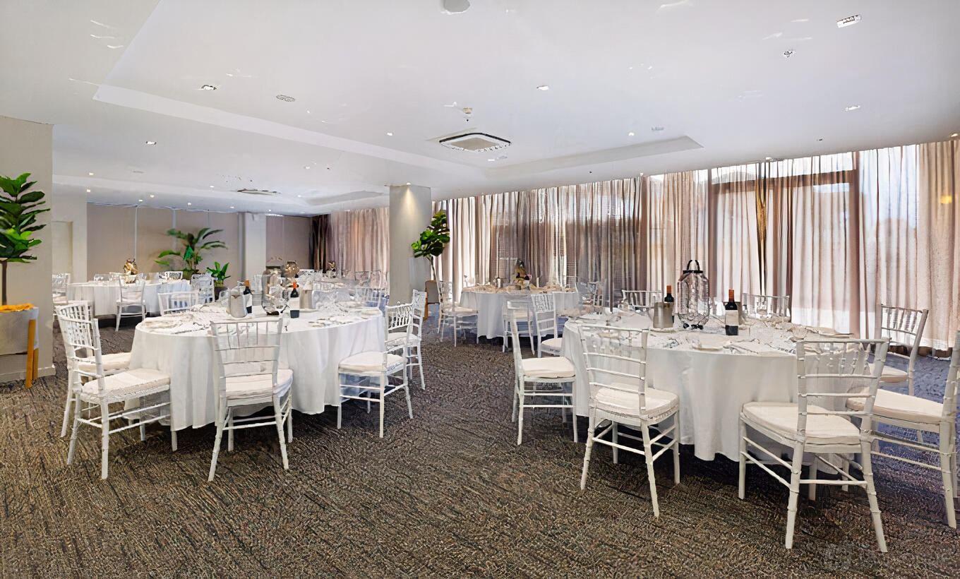 An event space with round tables dressed in white linens, white chairs, and floor-to-ceiling sheer curtains at Sage Hotel Wollongong.