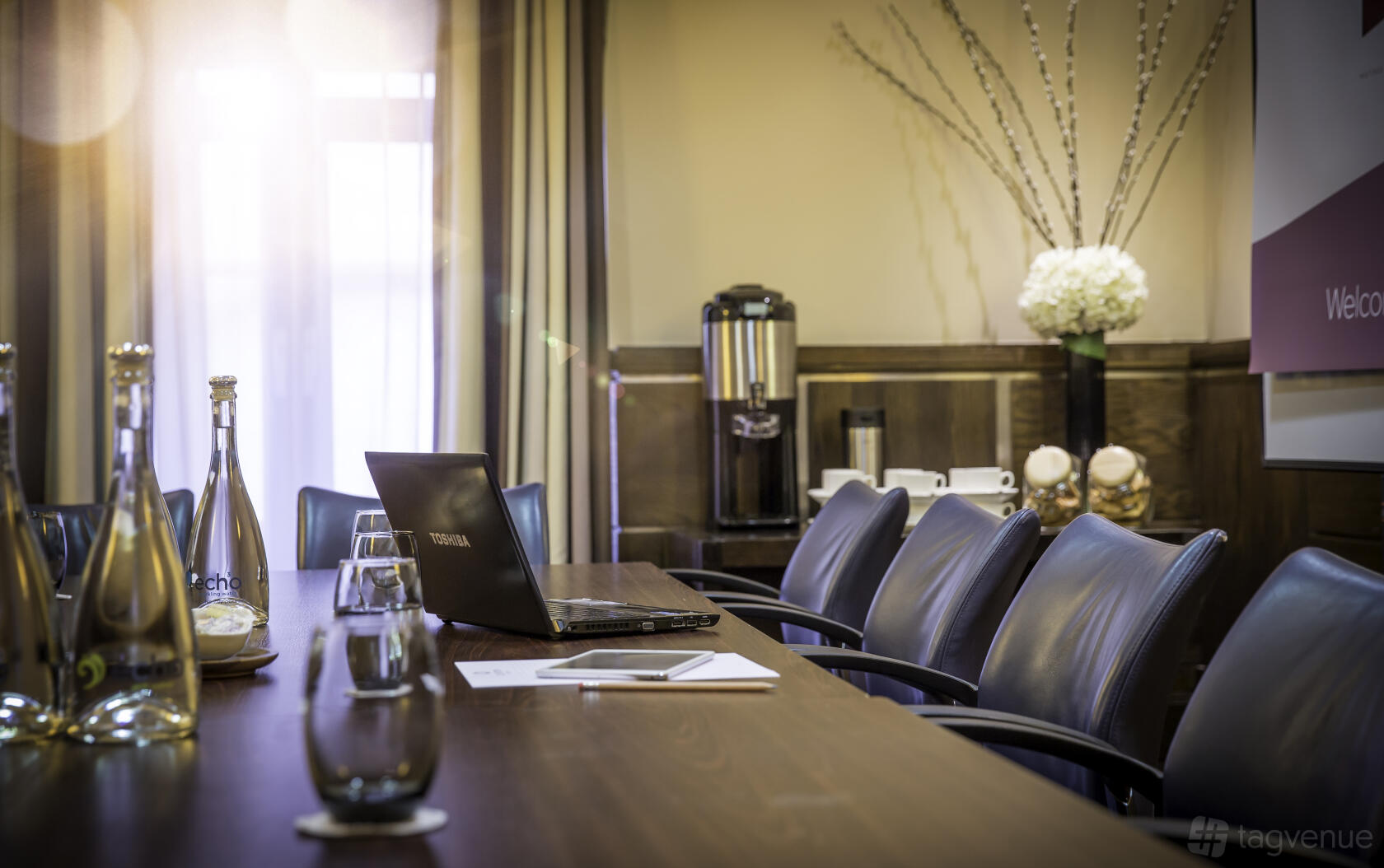 A meeting room with a long wooden table, leather chairs, a laptop, bottled water, and a coffee station at The Crown London Hotel.