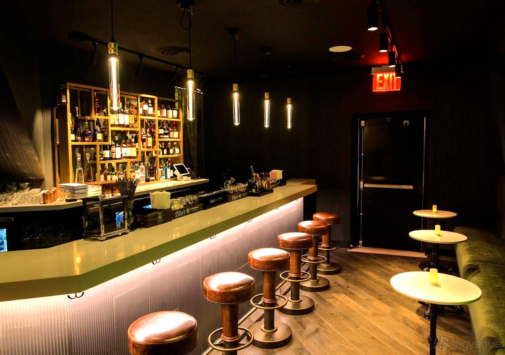 An event space in a bar with a stocked backlit counter, round bar stools, pendant lights, and small candlelit tables at Infamous.