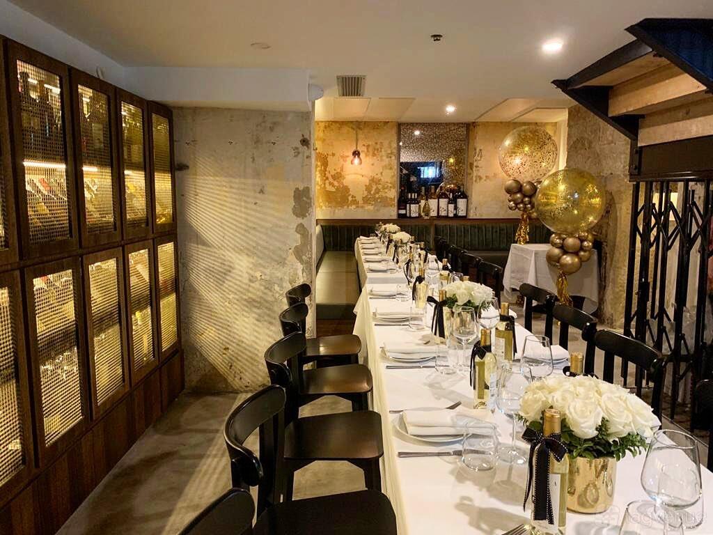 A restaurant with a long table set for dining, black chairs, floral centerpieces, and a wall wine display at Figo Ristorante & Bar.