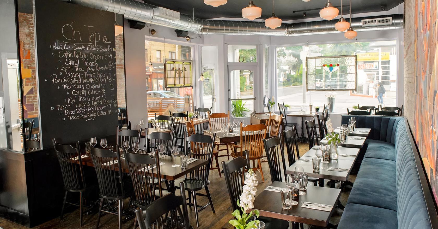 A restaurant with floor-to-ceiling windows, wooden tables set with glassware, and a chalkboard beer menu at Her Father's Cider Bar + Kitchen.