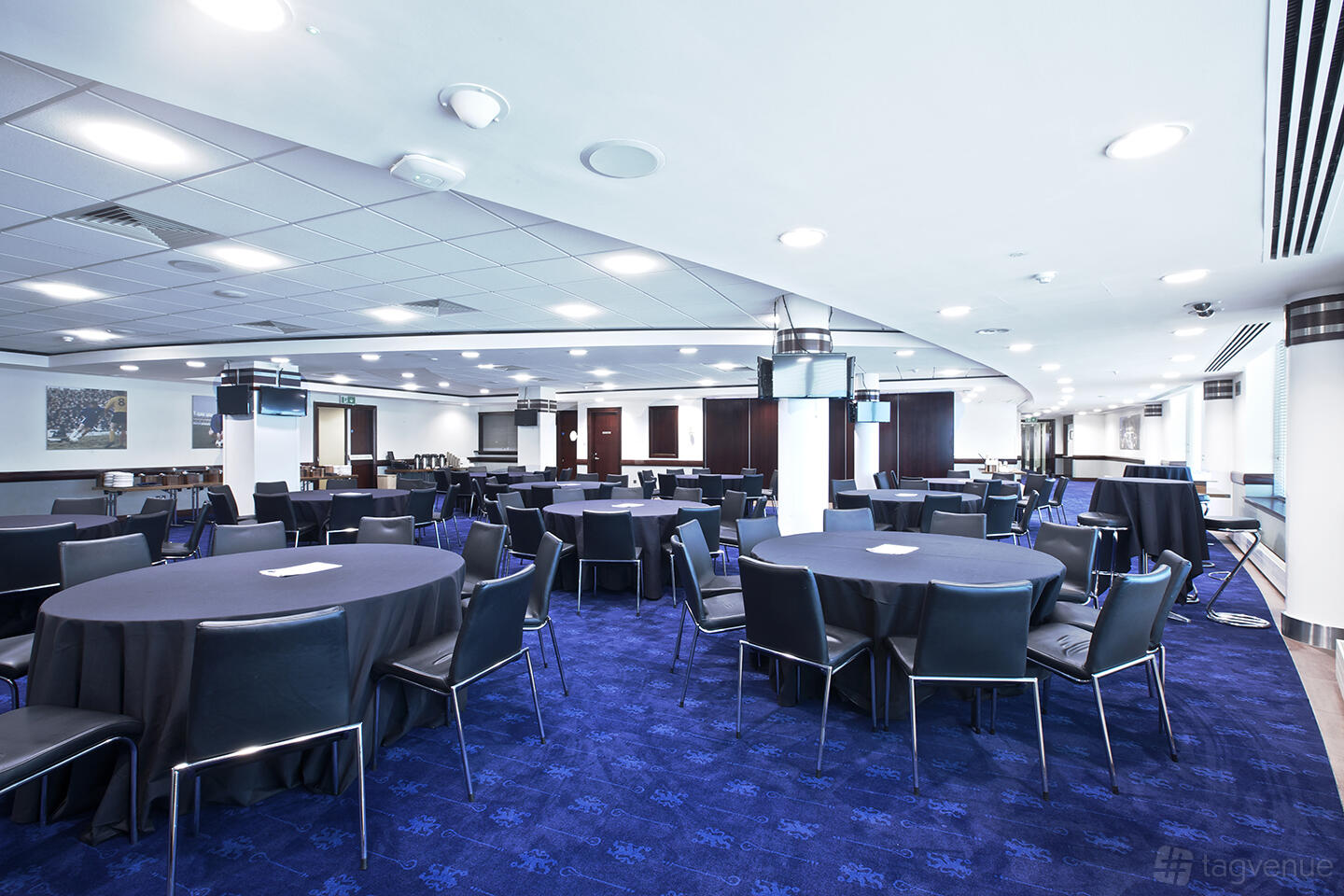 An event venue with round tables, black chairs, and blue carpet at Chelsea Football Club.