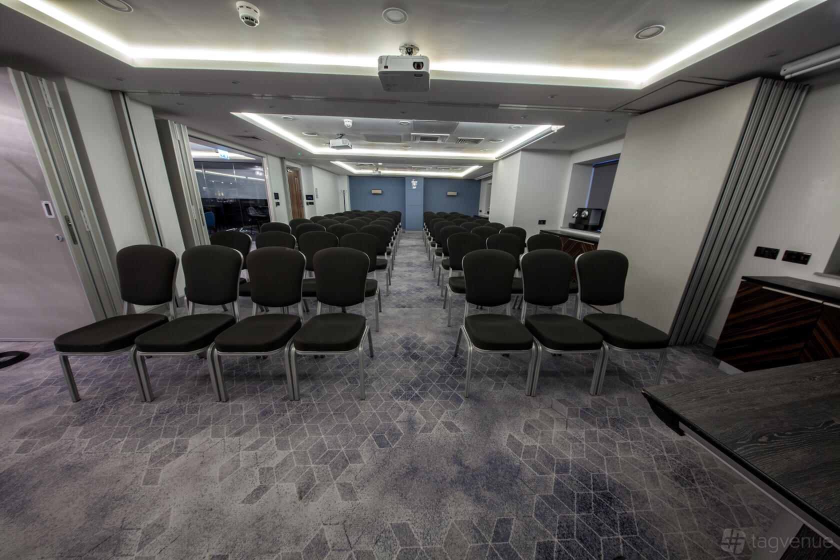 A hotel meeting room with rows of black chairs, patterned carpet, and ceiling projectors at The BCEC.