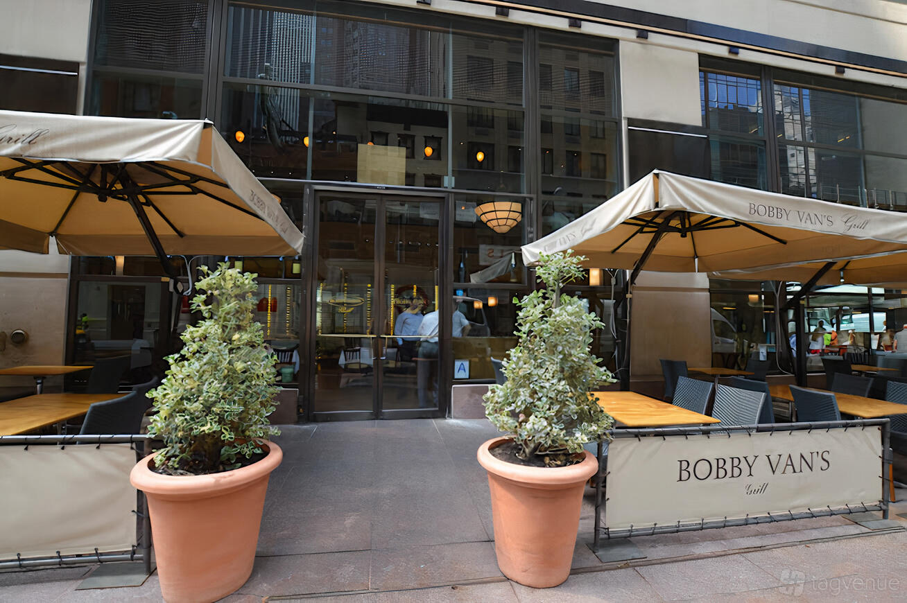 A restaurant with a sidewalk patio, tan umbrellas, potted plants, and outdoor dining tables at Bobby Van's (45th Street).