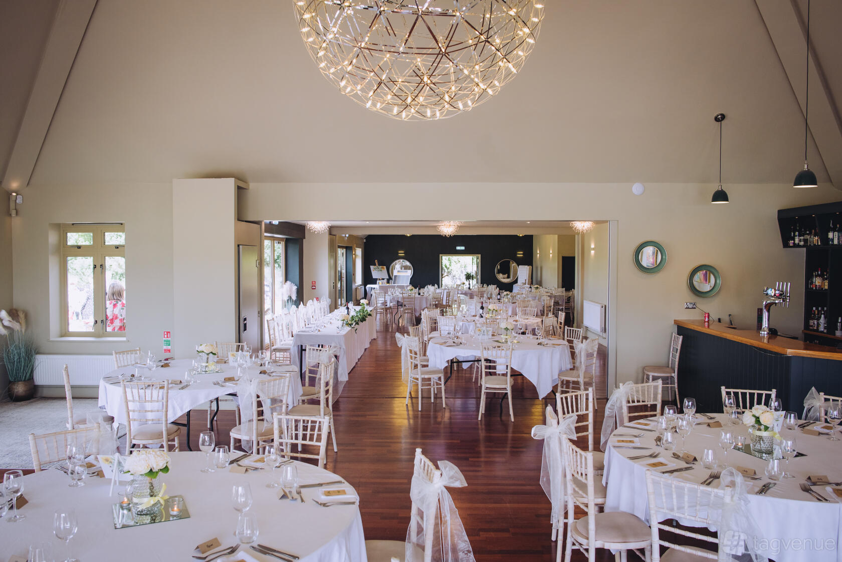 A function room with round tables draped in white linens, wooden floors, and large windows at The Club at Mill Green.