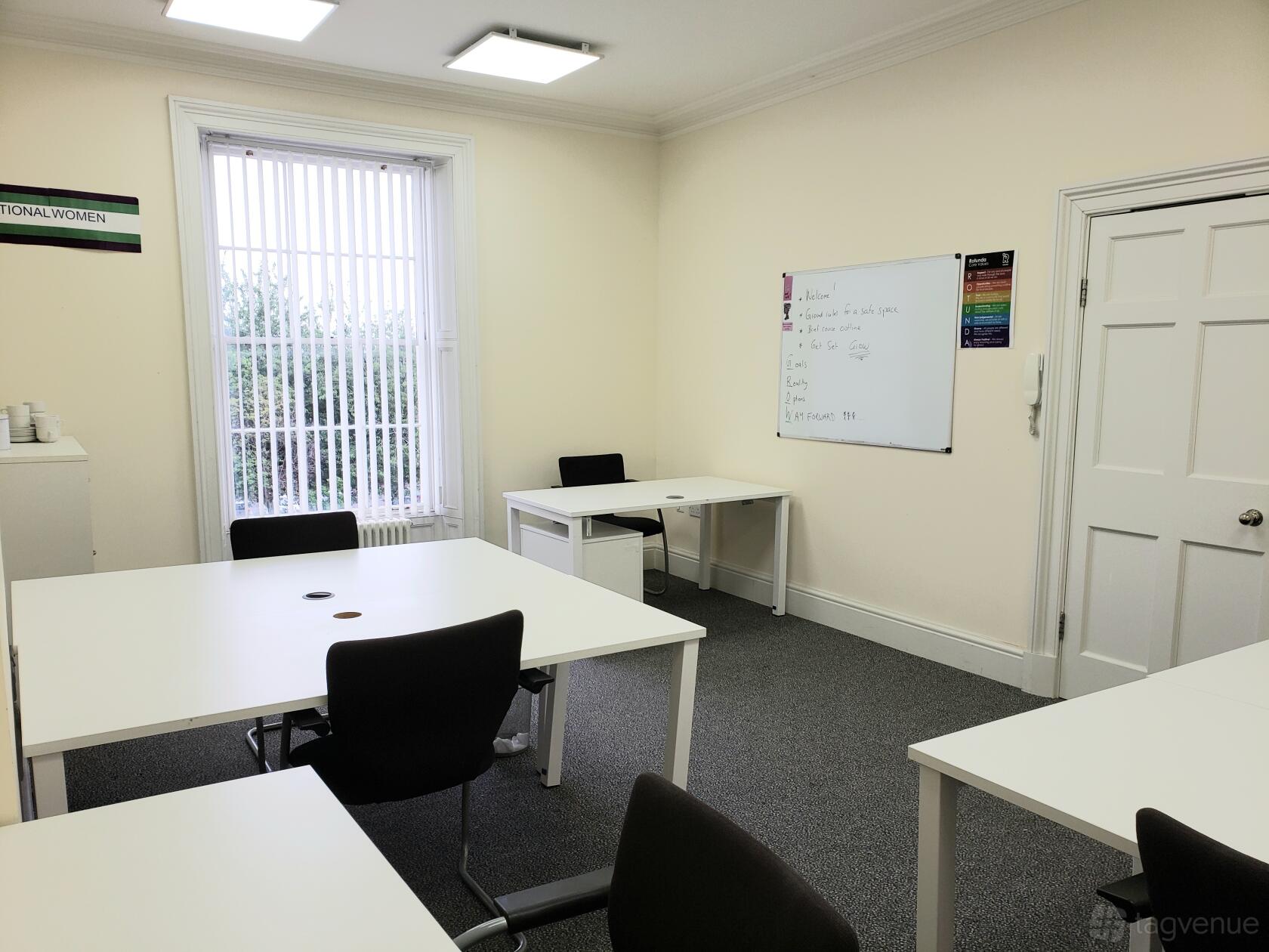 An office space with white desks, black chairs, large window with vertical blinds, and a wall-mounted whiteboard at The Rotunda.