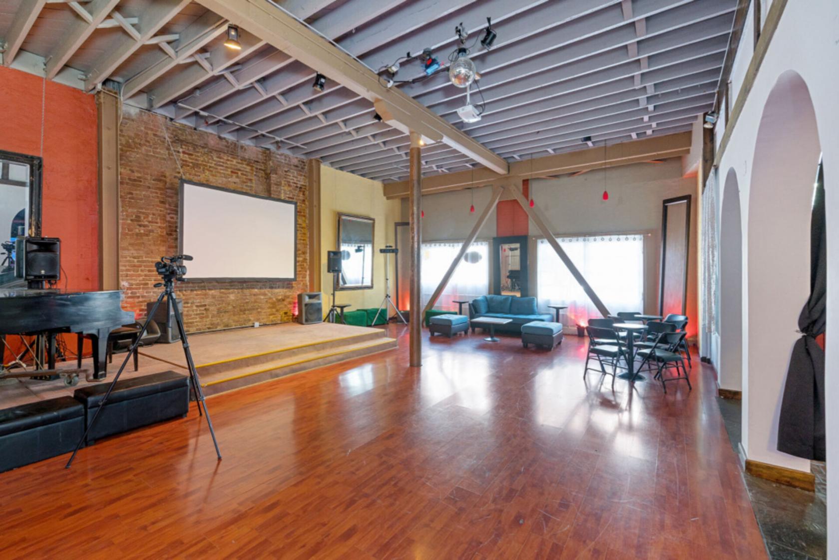 A lounge with exposed brick walls, a small stage, hardwood floors, and seating areas at Fame Venue.
