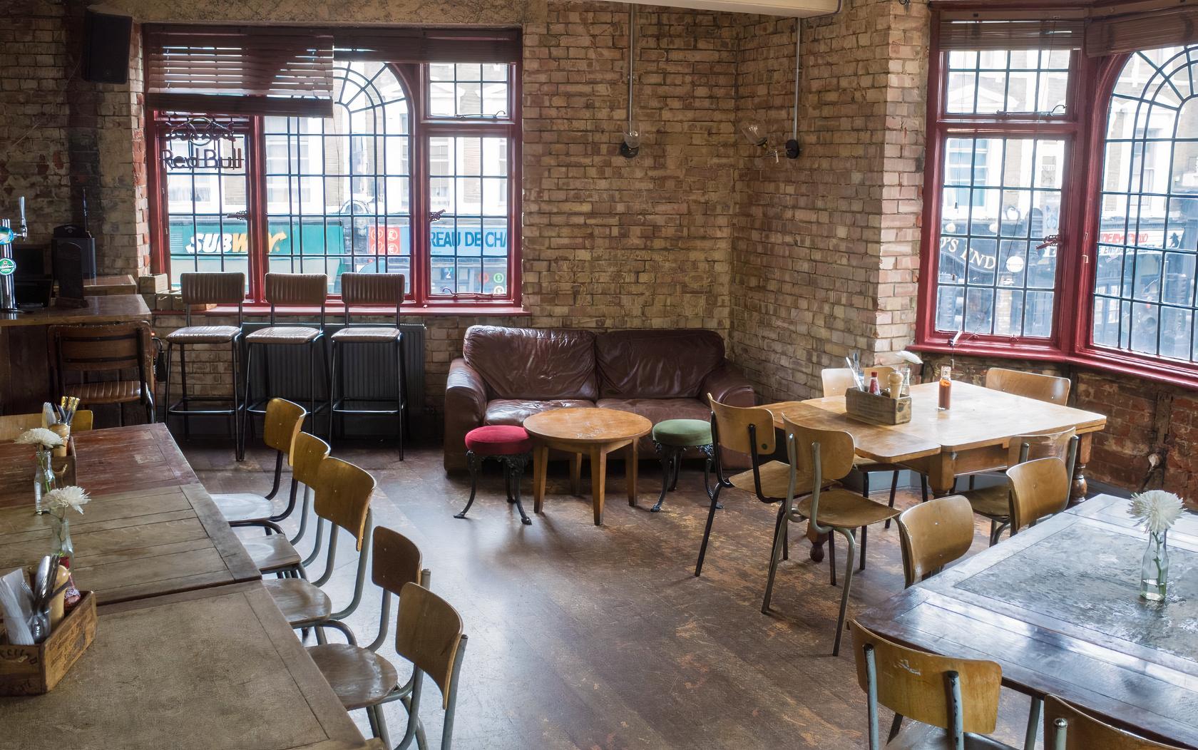 A function room in a pub with exposed brick walls, large arched windows, and mixed wooden tables at The Camden Eye.