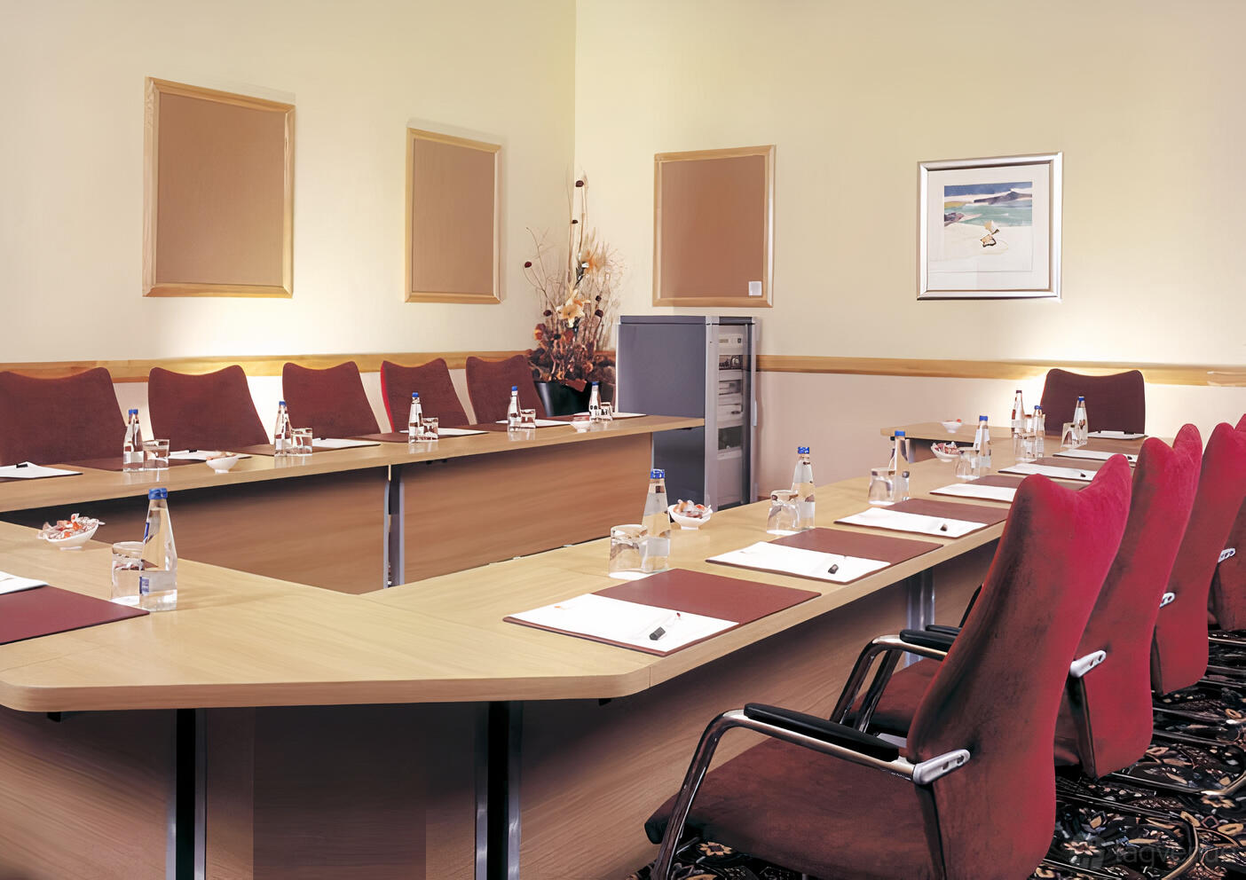 A hotel meeting room with a U-shaped conference table, red upholstered chairs, notepads, and bottled water at Grand Hotel.