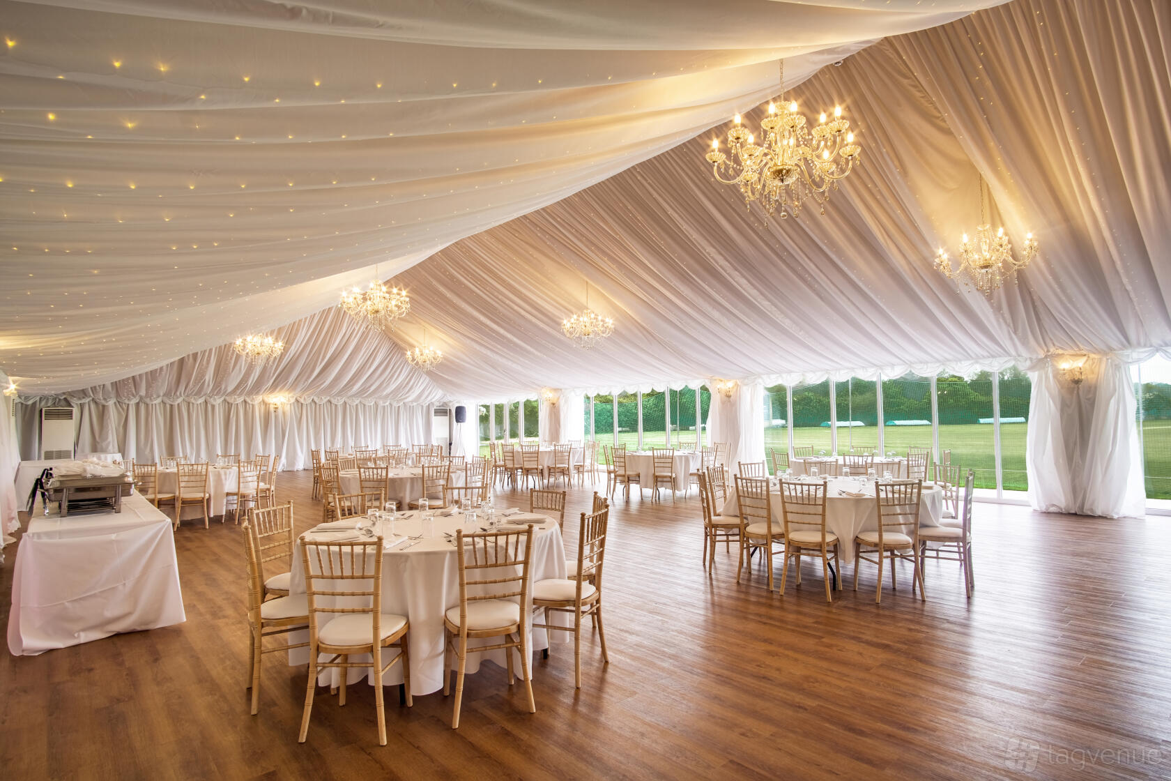 A hall with draped ceilings, chandeliers, wooden flooring, and round tables set by large windows at London Shenley Club.