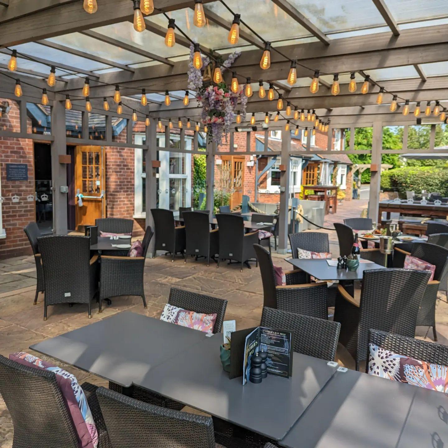 A covered garden seating area with rattan chairs, string lights, and stone flooring at The Crown.