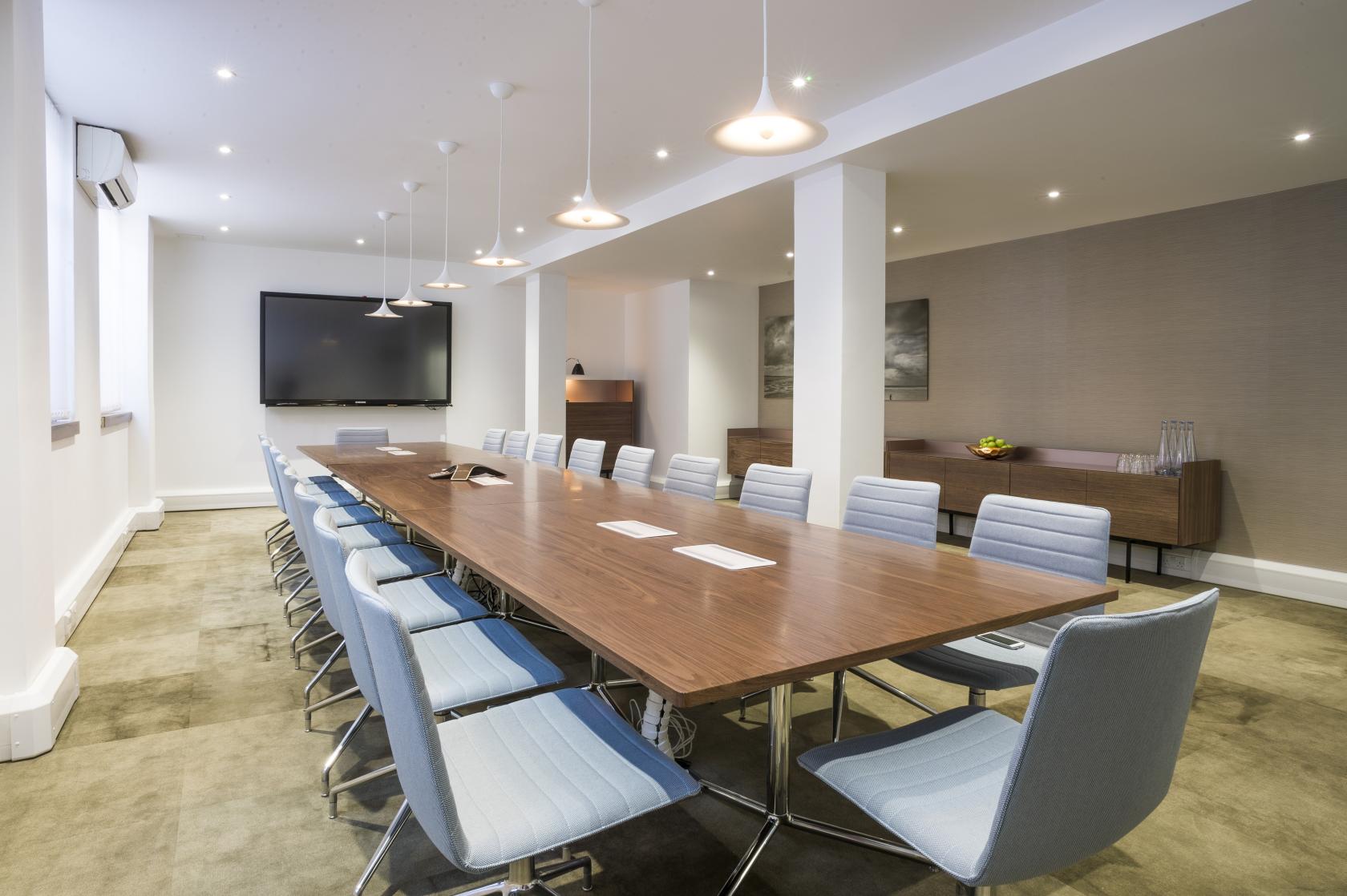 A boardroom with a long wooden conference table, modern blue chairs, pendant lights, and a wall-mounted screen at The Derby.