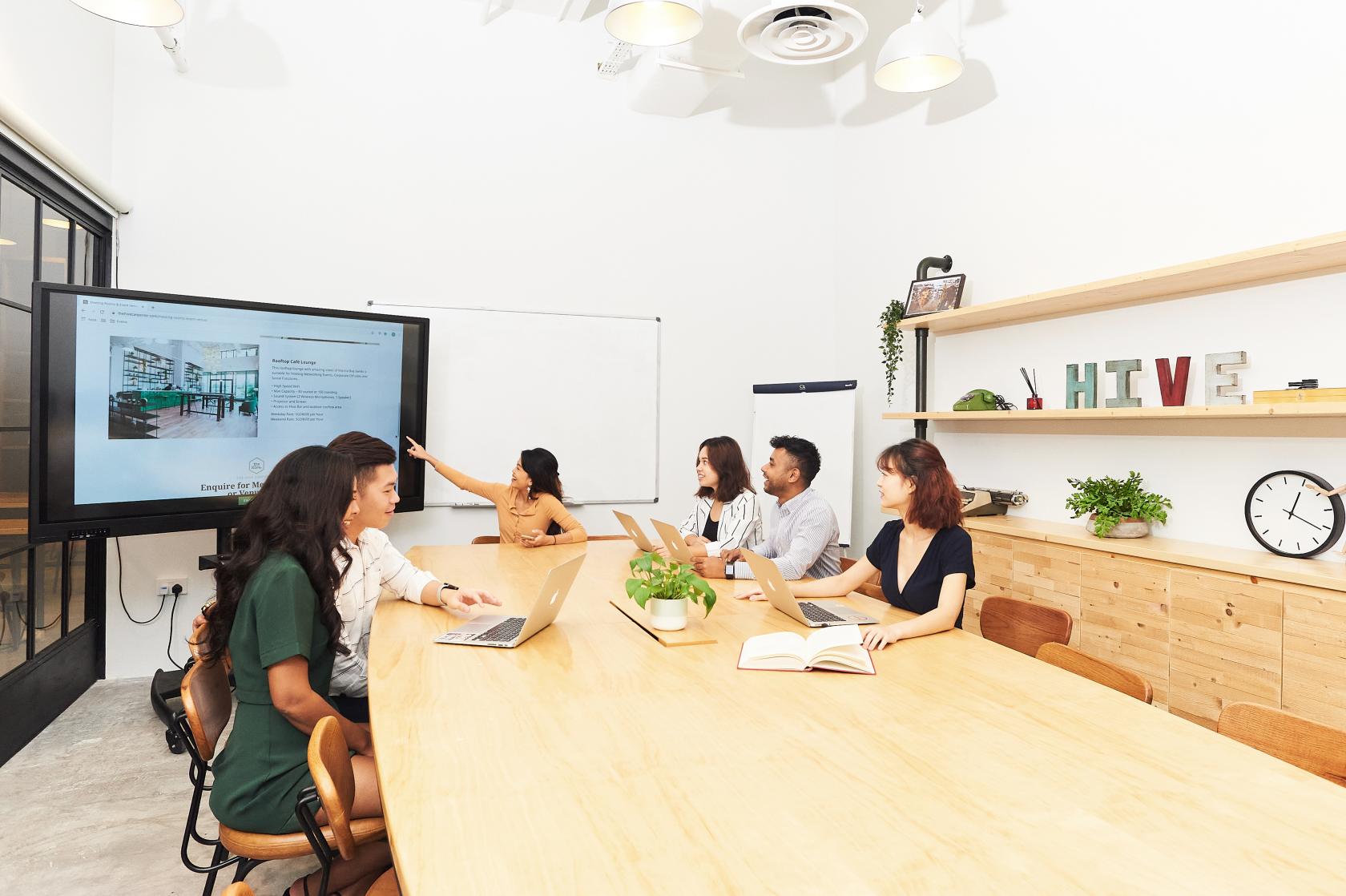 A coworking meeting room with a long wooden table, wall-mounted screen, and open shelving at The Hive Carpenter.
