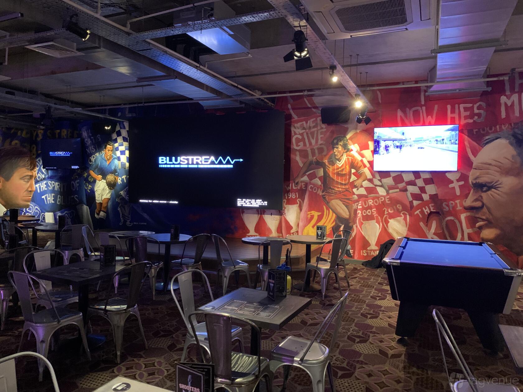 A bar with sports-themed mural walls, a large screen, and metal tables with chairs at The Bierkeller Liverpool.
