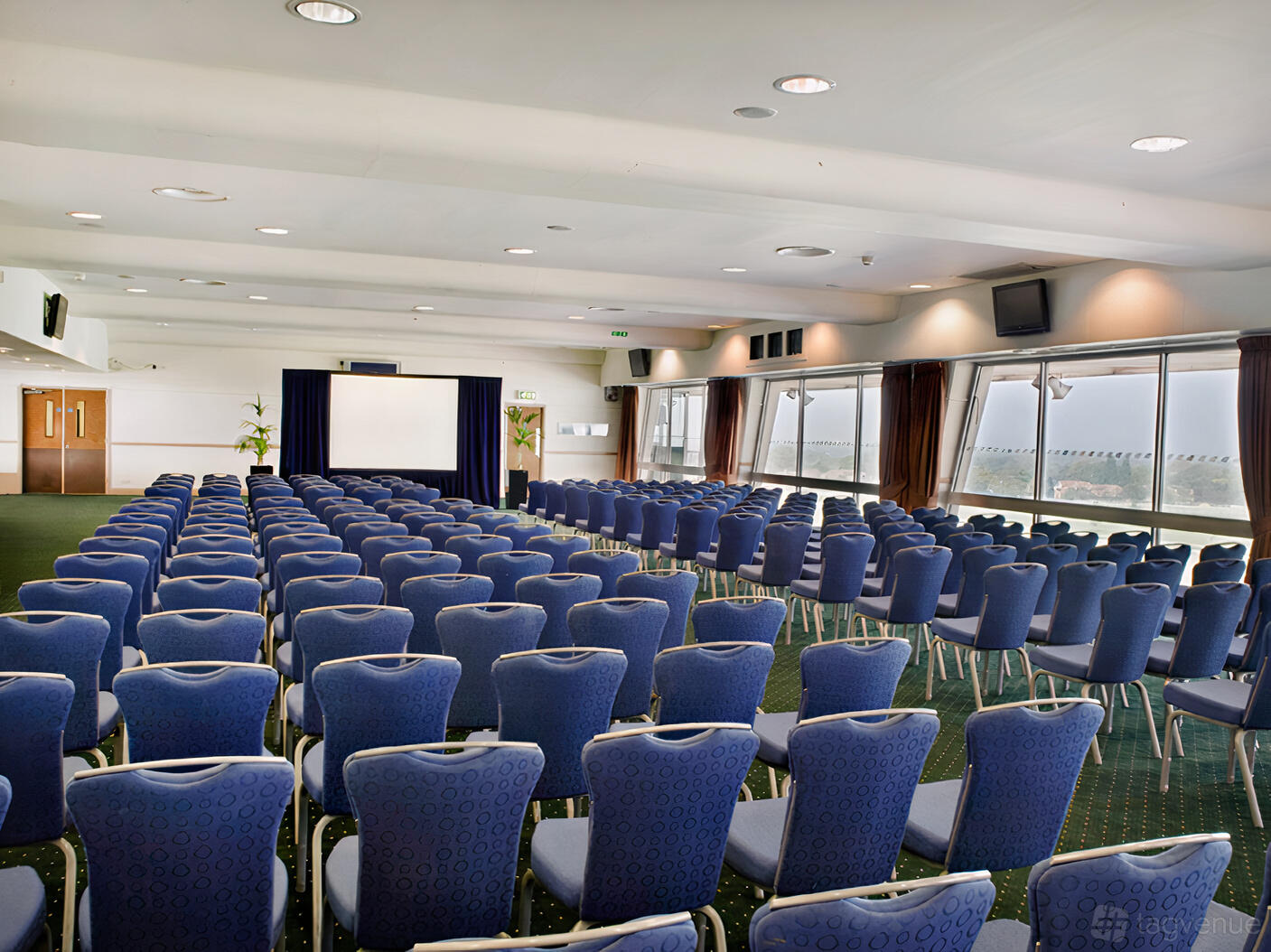 An event venue with rows of blue chairs facing a projector screen, large windows, and carpeted floors at Sandown Park Racecourse.