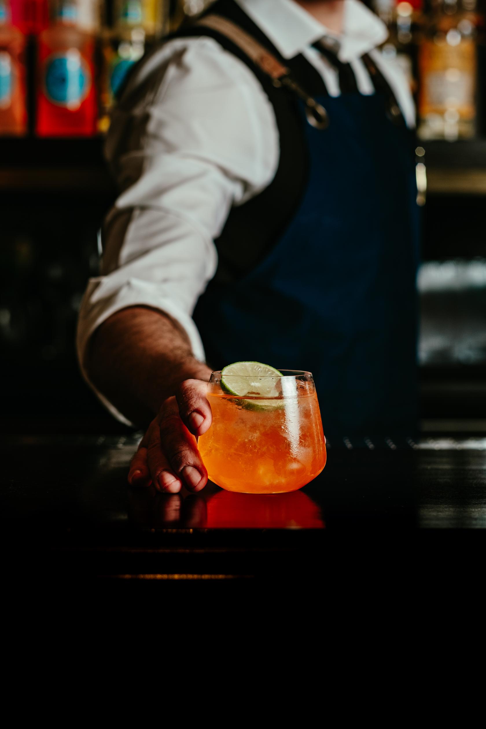 A cocktail bar with a bartender serving an orange cocktail garnished with lime at LOCKES Bar Covent Garden.