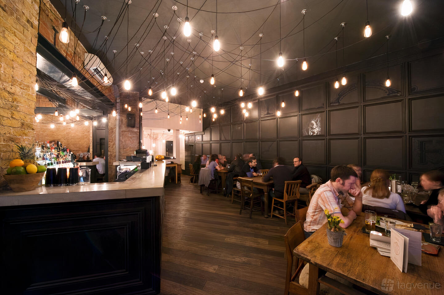 A bar with exposed brick, dark wood floors, pendant lights, and casual seating at The Alice House West Hampstead.