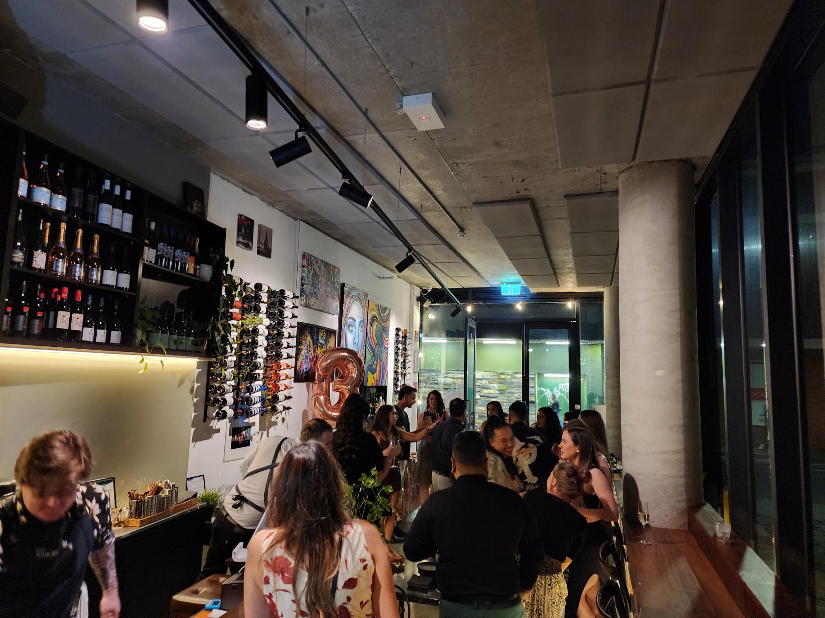 A bar with floor-to-ceiling windows, concrete ceiling, wall-mounted wine racks, and guests seated at Ruelle Wine Bar.