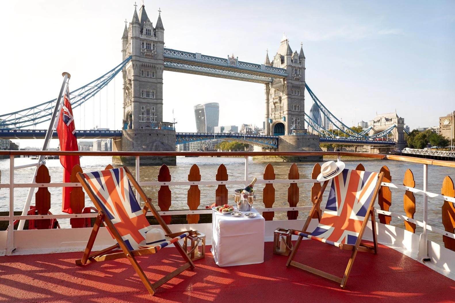 A boat deck with Union Jack deck chairs, a table set for tea, and Tower Bridge in view on Elizabethan – Thames Luxury Charters.