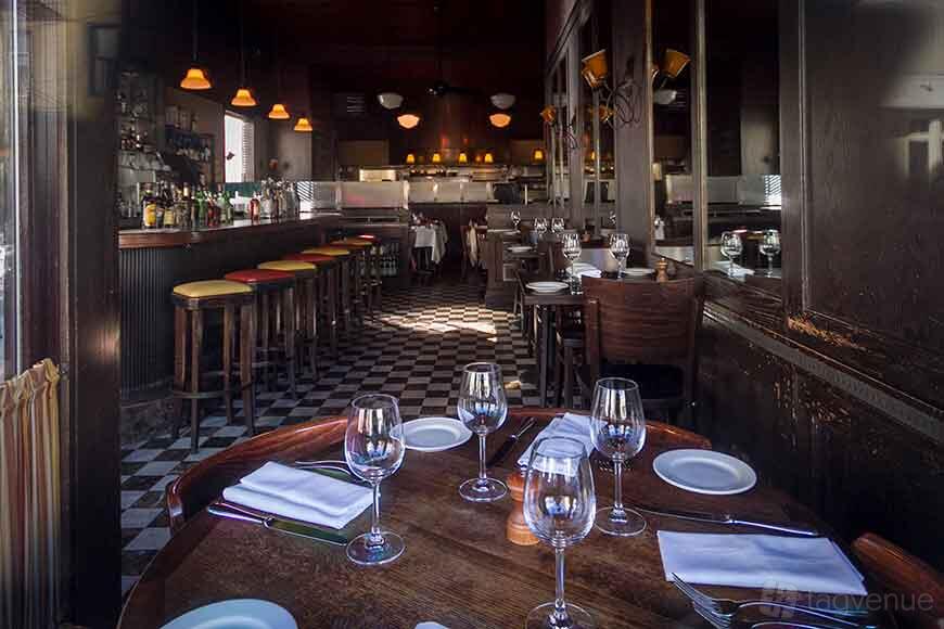 A restaurant dining area with checkered floors, wood tables set with glassware, and a bar with colorful stools at Florio.