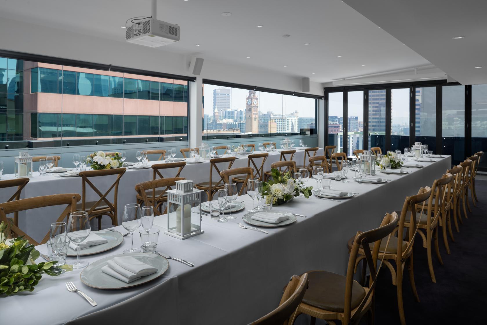 An event space with floor-to-ceiling windows, city views, and long banquet tables set for dining at Rydges Sydney Central.