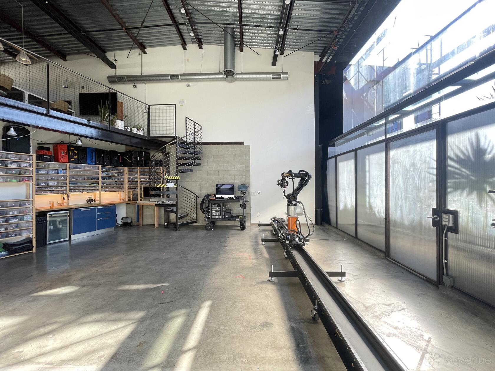 A photo studio with a robotic camera arm, concrete floors, wall-mounted tool shelves, and floor-to-ceiling windows at Ms Robot.