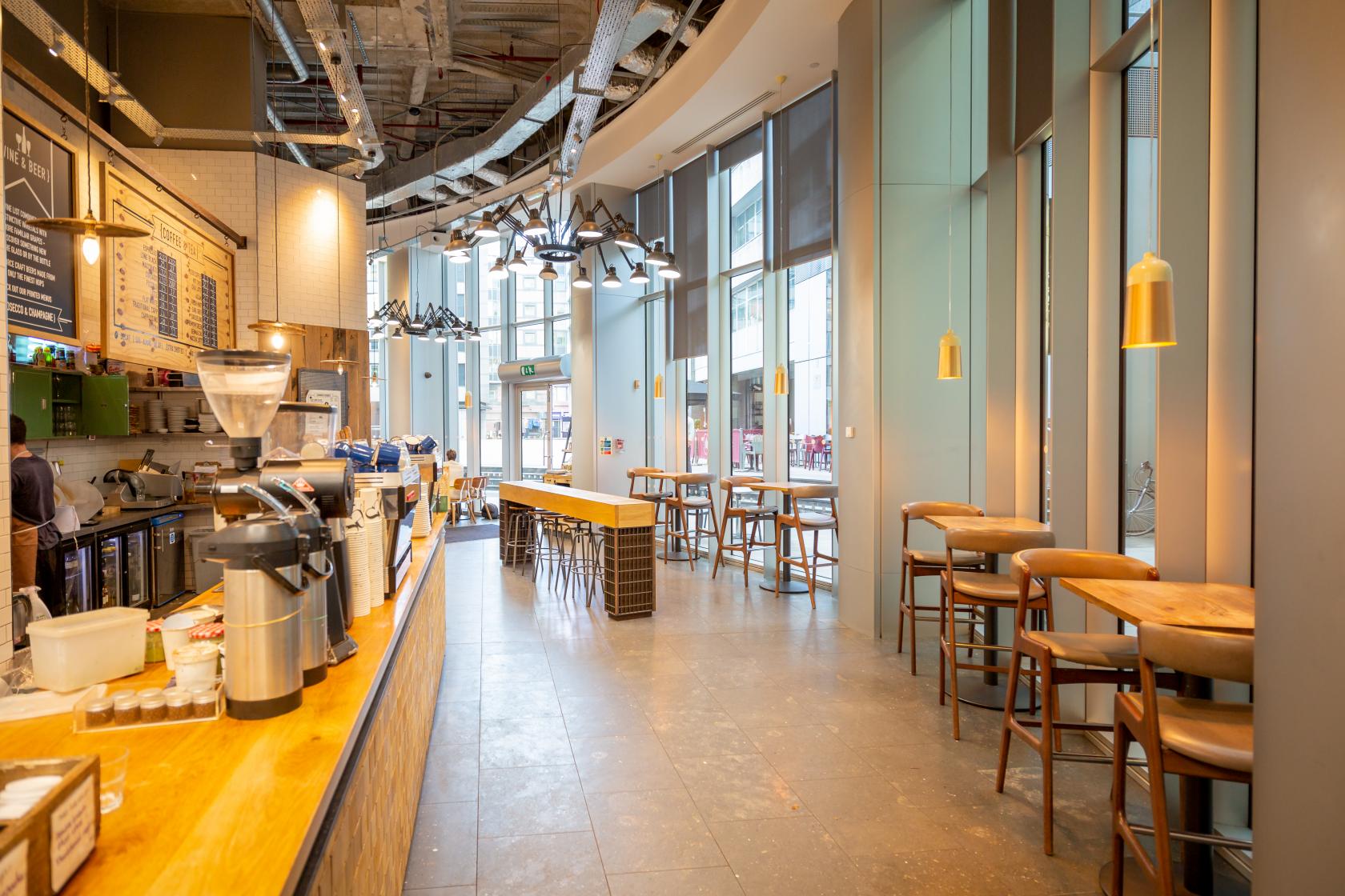 A cafe with floor-to-ceiling windows, high ceilings, and communal wooden tables at Notes Coffee & Bars - Moorgate.