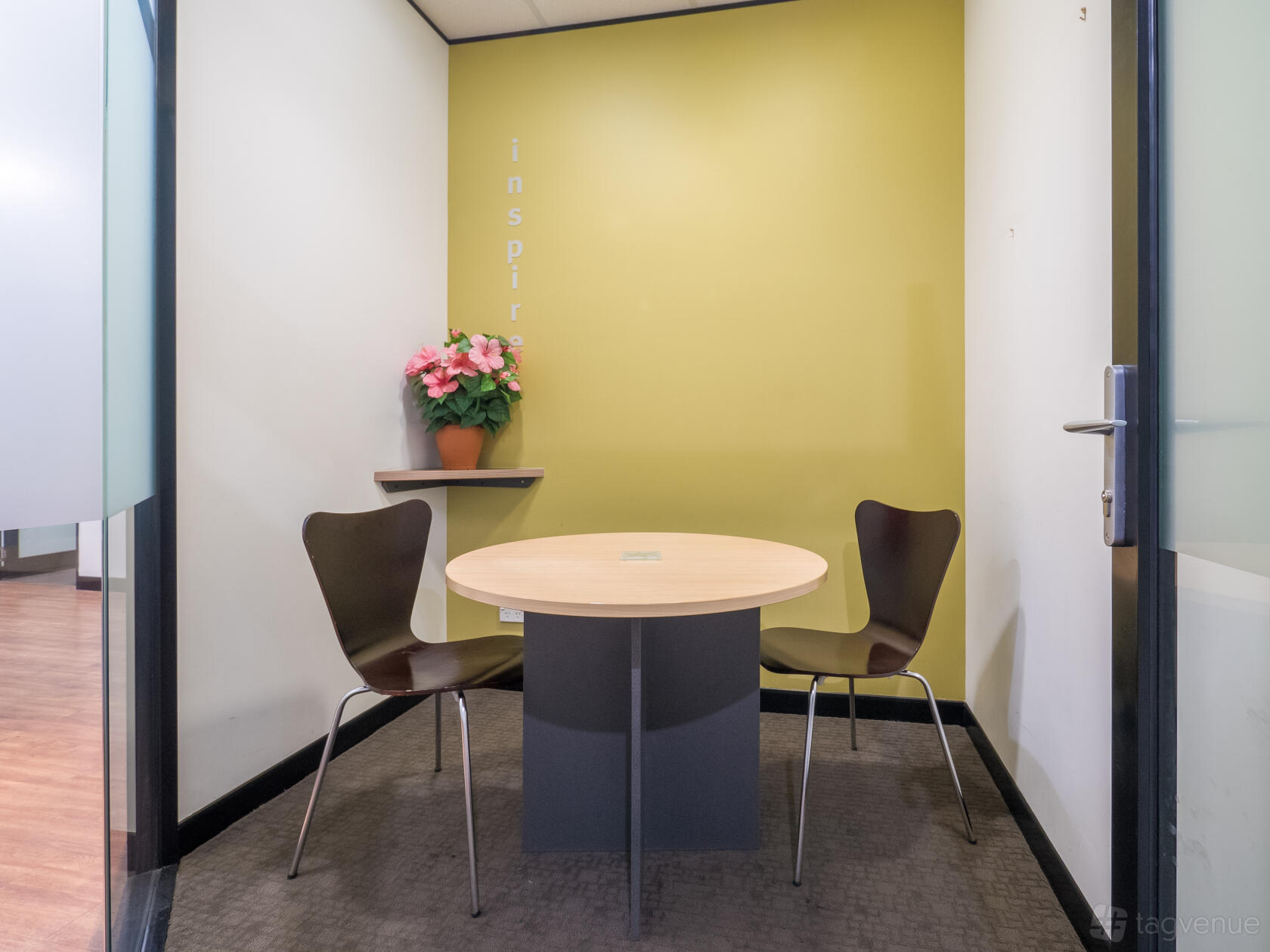 A coworking meeting room with two chairs, a round table, and a wall shelf with a potted plant at M1 Business Centre.