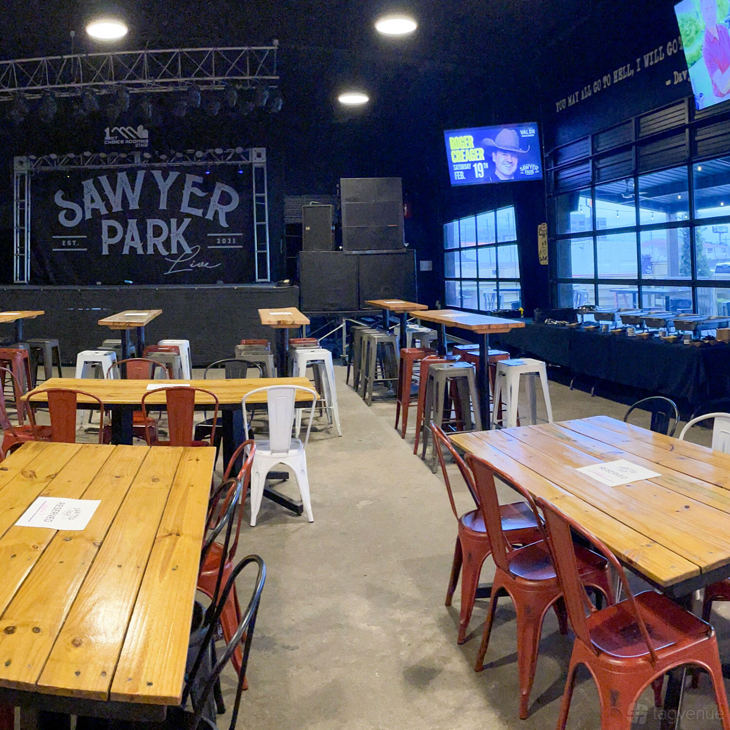 A bar with long wooden tables, metal chairs, a stage with speakers, and large windows at Sawyer Park Ice House.
