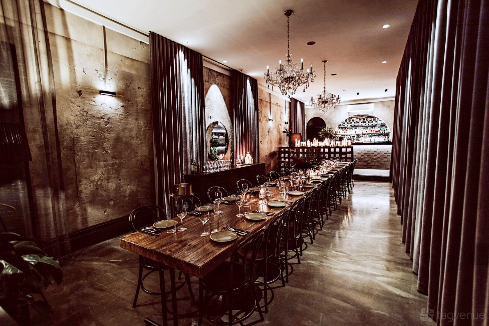A dining room with a long wooden table, chandeliers, velvet curtains, and exposed brick walls at Aunty Kim's.