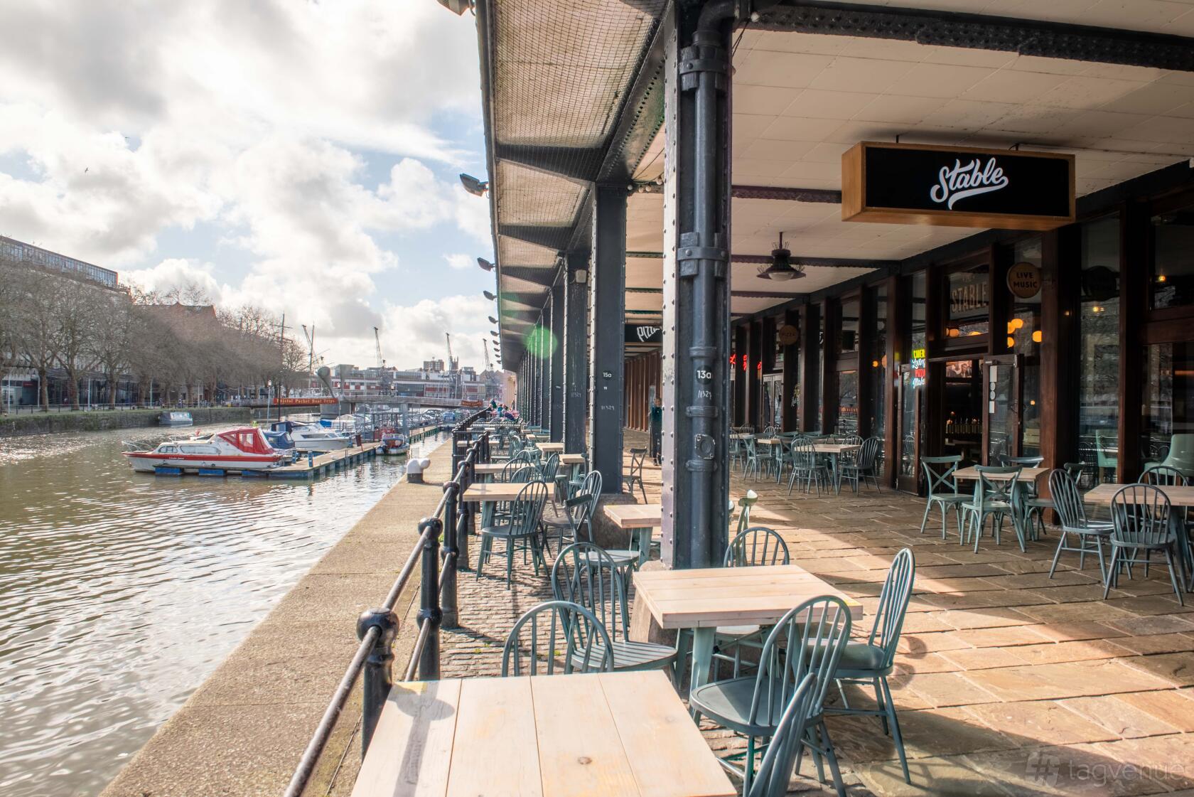 A pub with an outdoor riverside terrace, metal chairs, and wooden tables at The Bristol Stable.