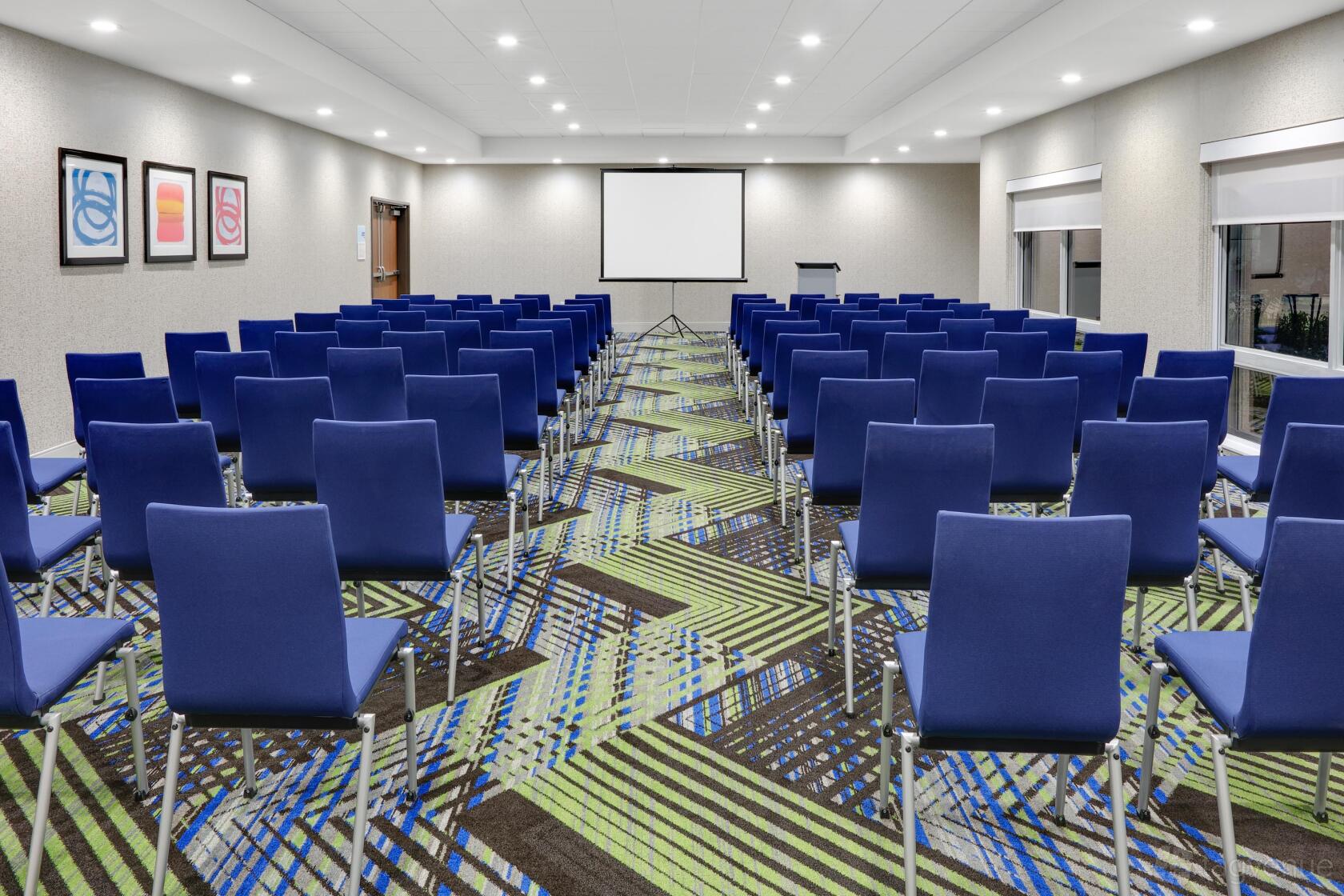 A banqueting hall with rows of blue chairs facing a projector screen and patterned carpet at Holiday Inn Express & Suites Dallas NW Hwy.