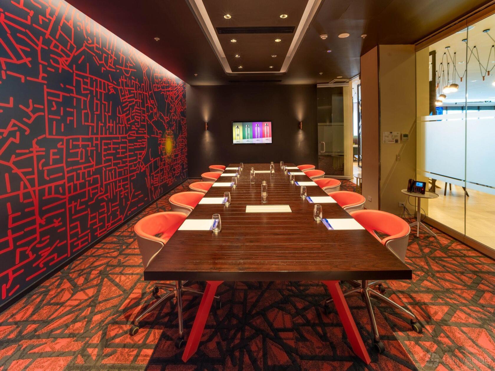 A meeting room with a long wooden table, red chairs, geometric wall art, and a mounted screen at Holiday Inn Express Adelaide City Centre.