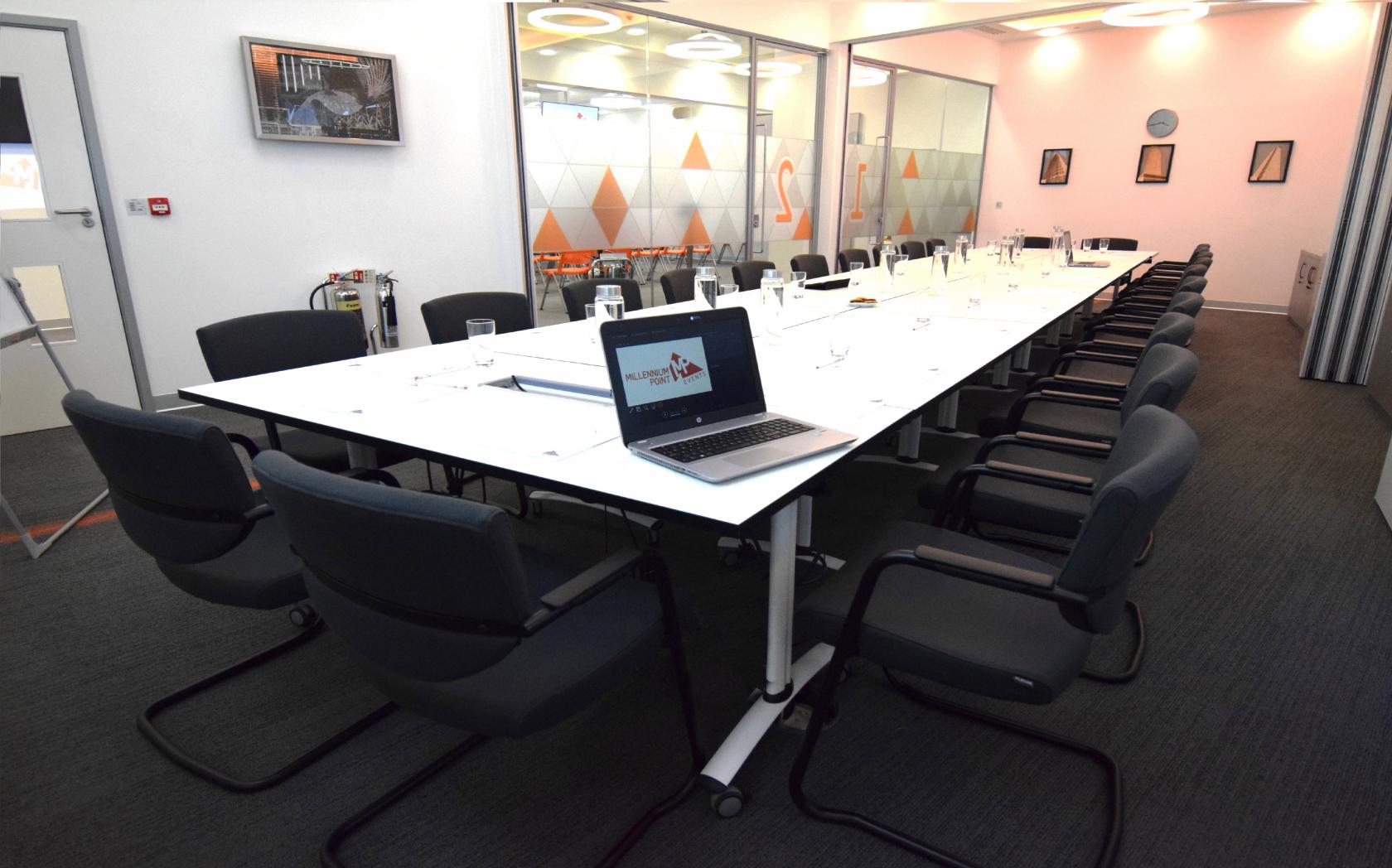 A boardroom with a long white conference table, black chairs, and glass walls with geometric frosted patterns at Millennium Point.