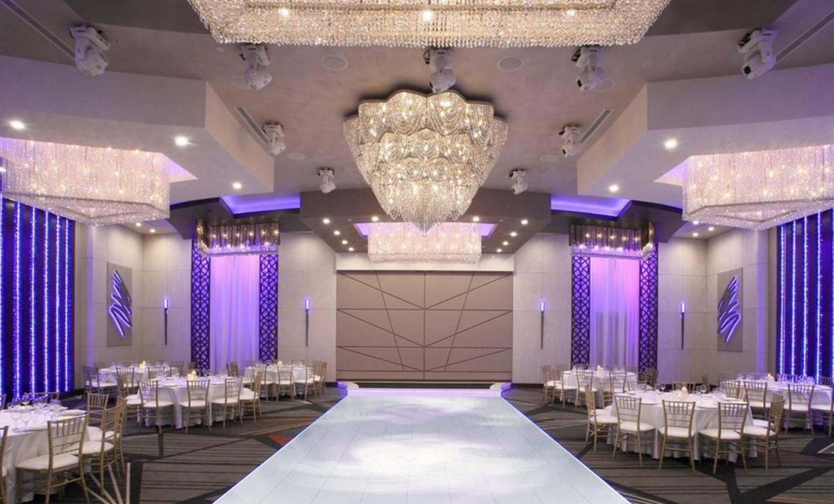 A ballroom with grand crystal chandeliers, uplighting, and round tables with white chairs at Allure Banquet & Catering.