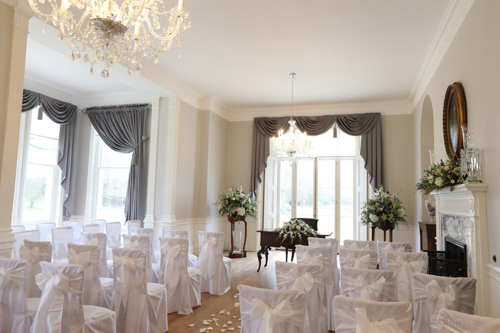 A country house ballroom with chandeliers, large windows, draped curtains, and chairs covered in white fabric at Hatfield Place.