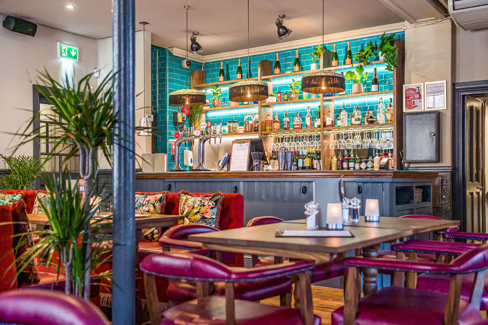 A lounge in a pub with teal tile backsplash, hanging lights, red leather chairs, and potted plants at The Crown & Two Chairmen.