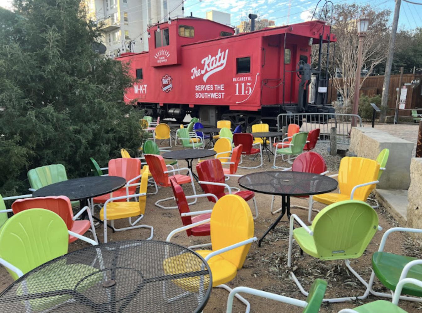 An outdoor terrace with colorful metal chairs and round tables beside a vintage red train car at Katy Trail Ice House.